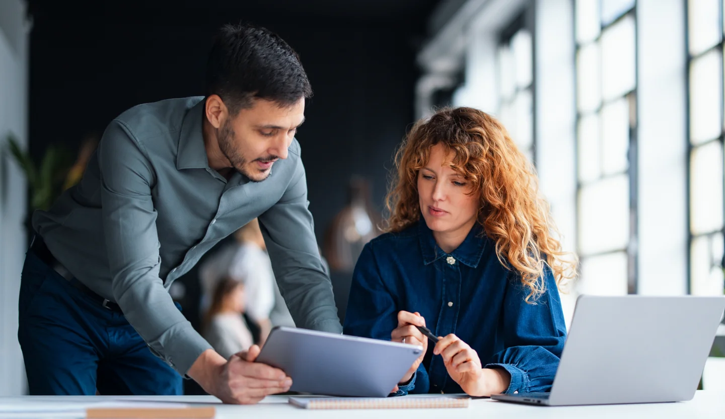 One office worker stands while another sits at a table, both of them looking at a tablet
