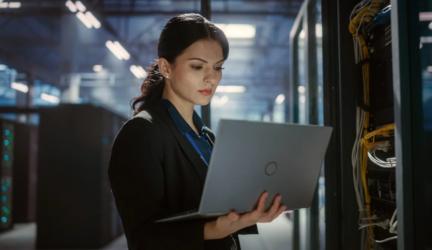 Woman stands in a server warehouse holding a laptop
