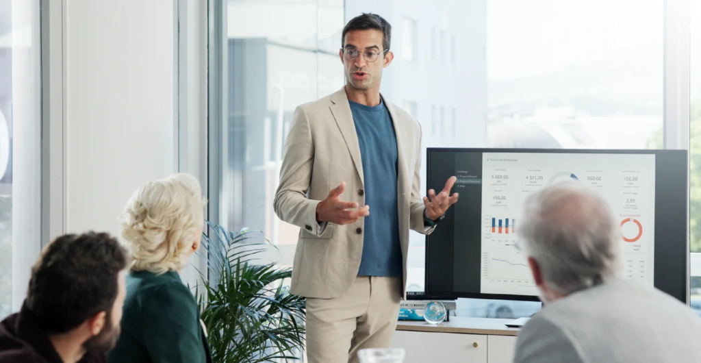 Office worker standing in front of a screen explaining something to people sitting around a table