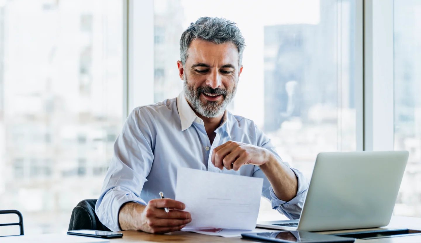Office worker sits at a desk in front of a laptop while holding a piece of paper