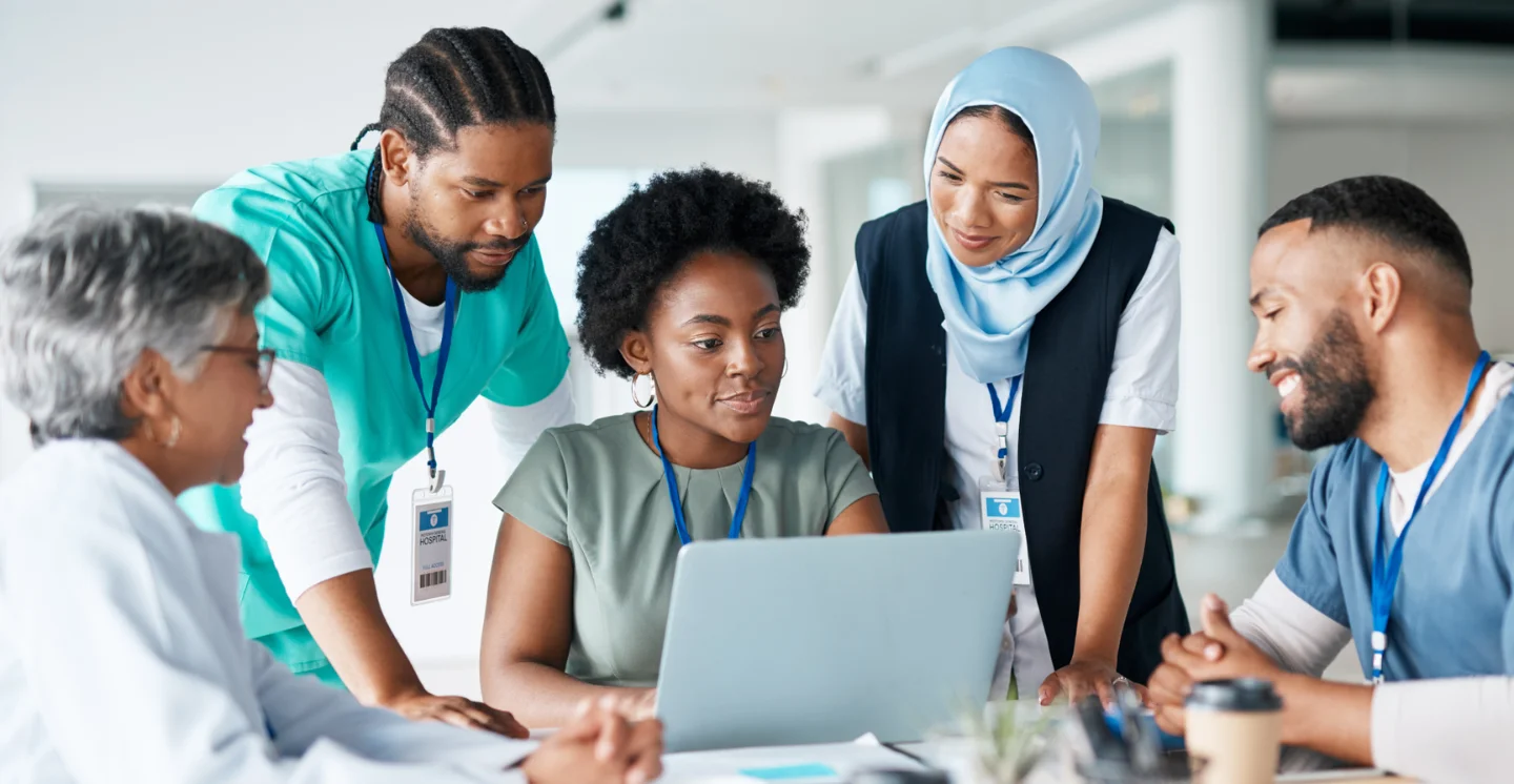 Three healthcare workers sit around a table with two more standing behind them, all looking at a laptop