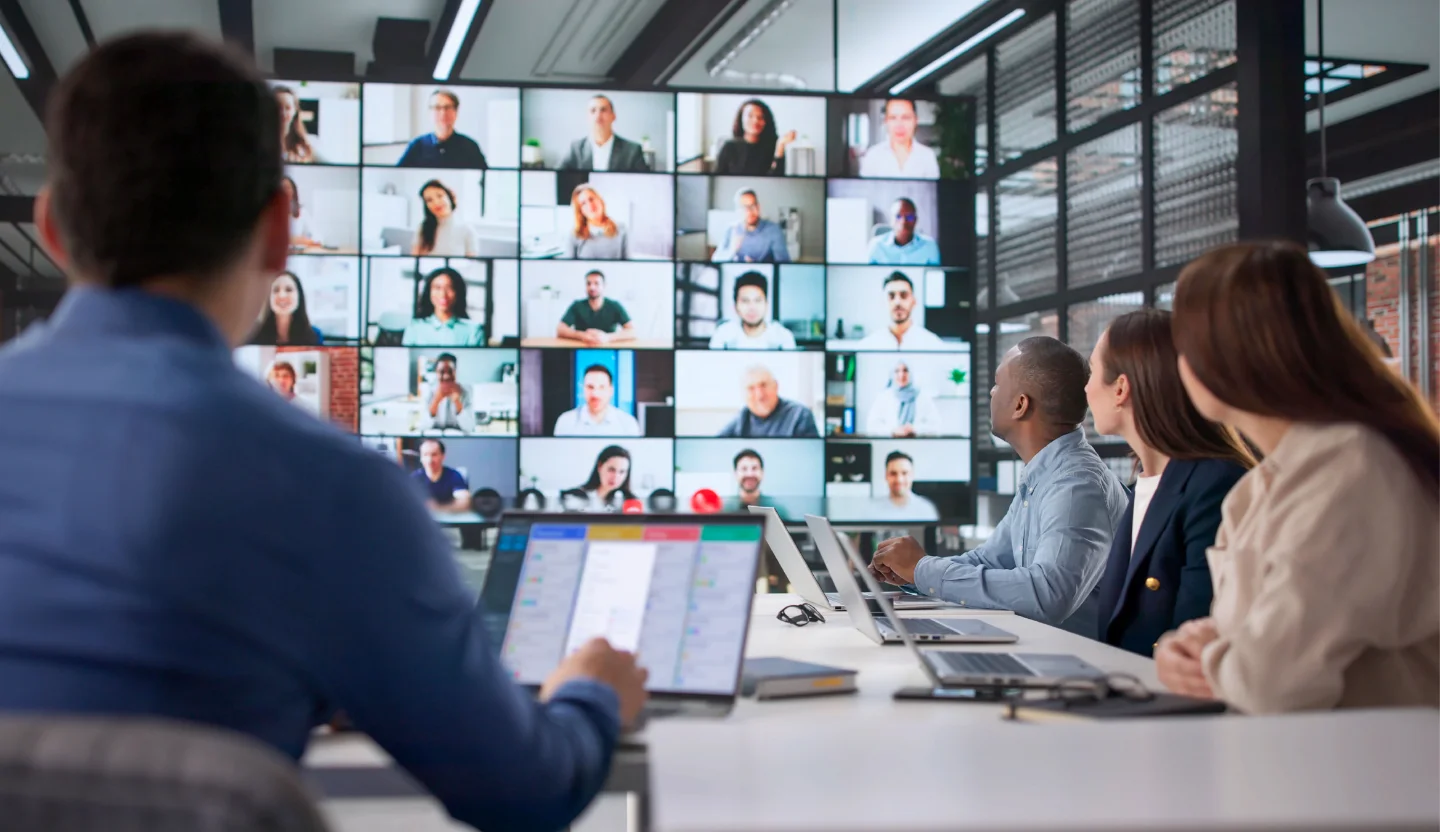 Four office workers sitting around a conference table looking at a large screen displaying several people on a video call