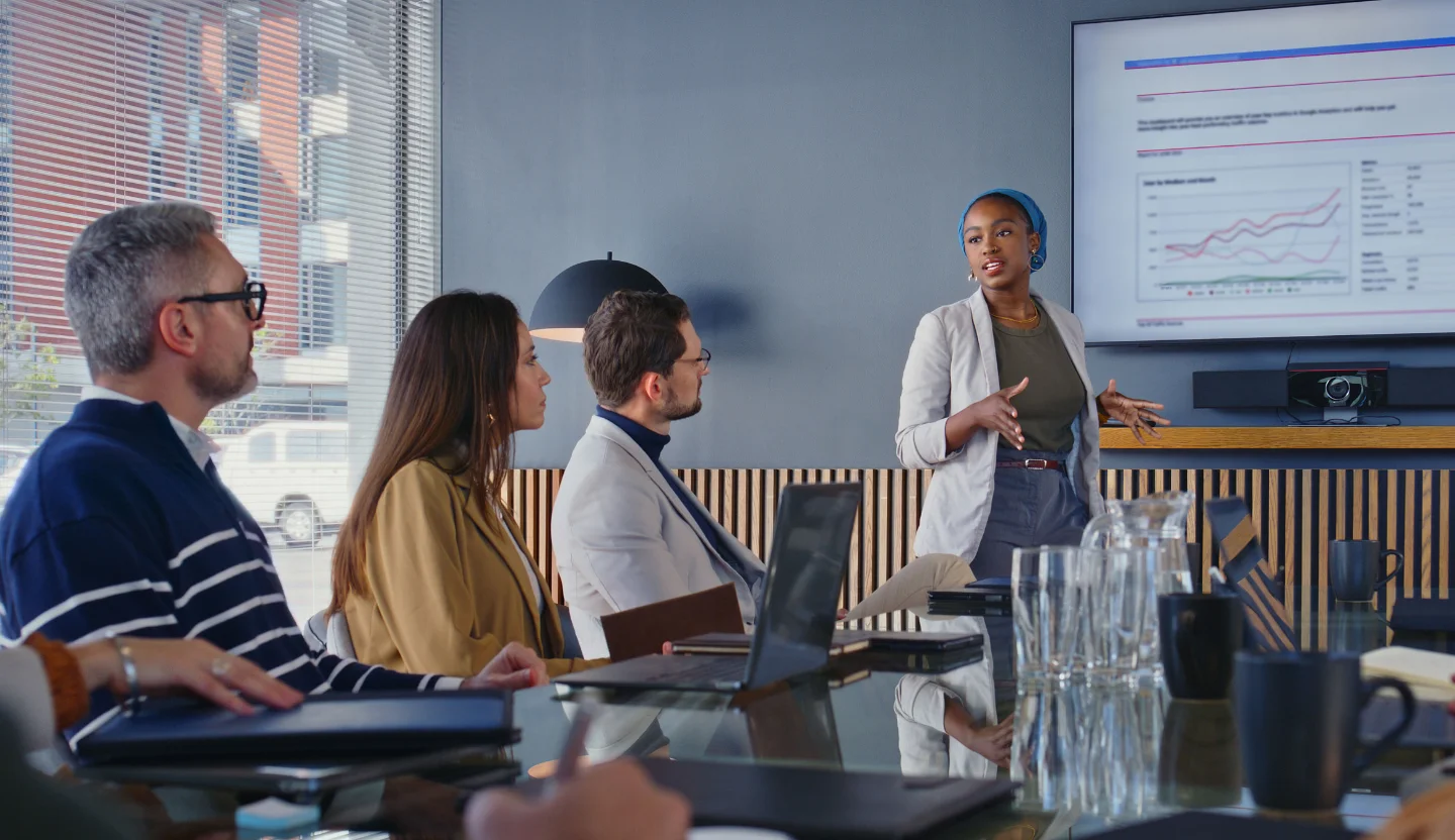 Three office workers sit at a table watching another person present in front of a screen