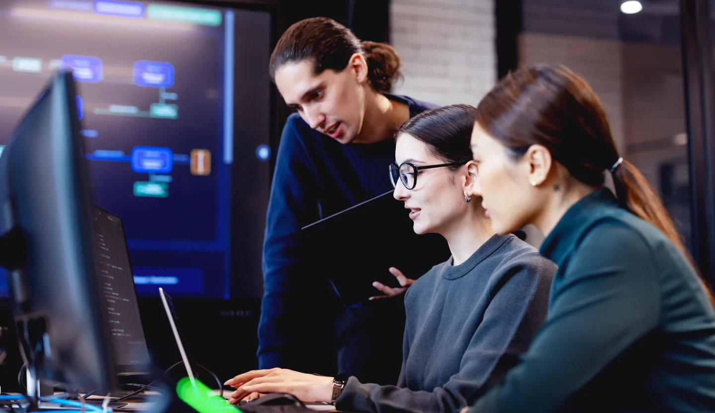 Two office workers sit in front of a computer while another person stands near them while holding a laptop