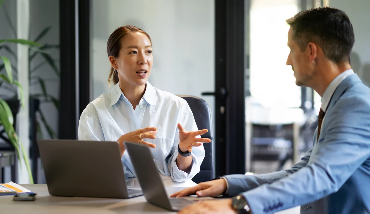 Two office workers have a discussion at a conference table with laptops in front of each of them