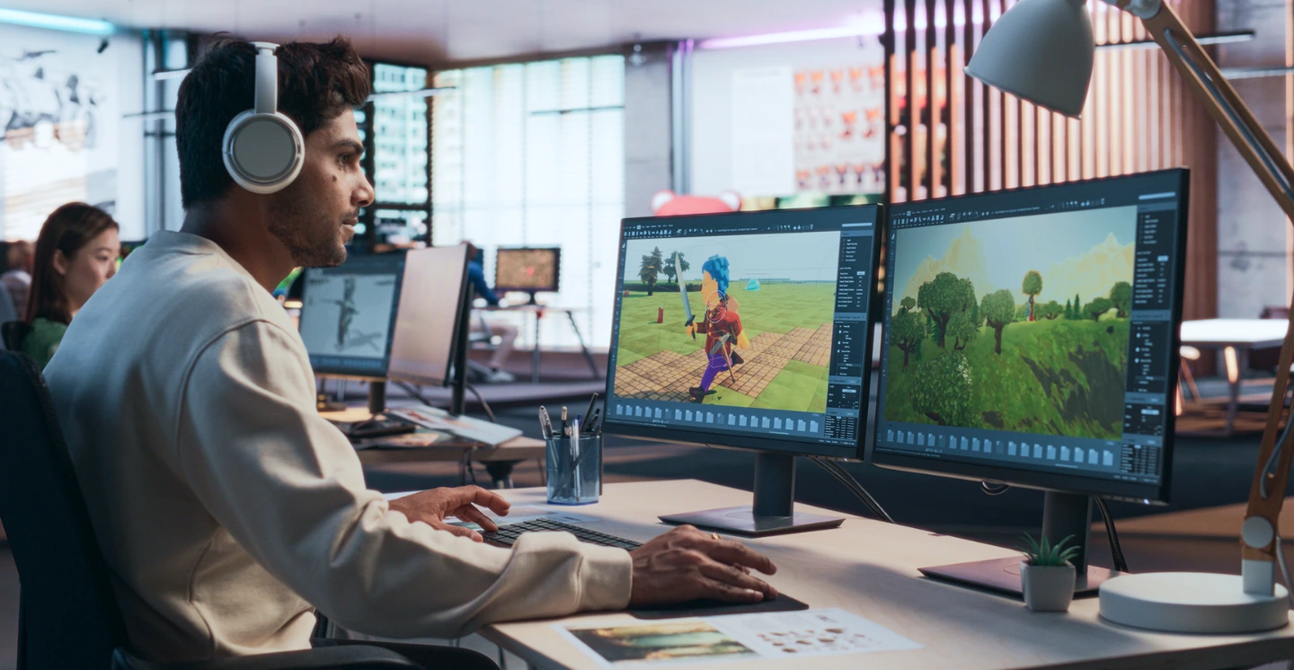 A video game developer works in front of two monitors while a coworker sits and works in the background